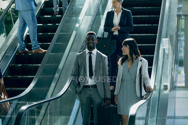 Vue de face de divers hommes d'affaires interagissant les uns avec les autres tout en utilisant des escaliers mécaniques dans le bureau moderne — Photo de stock