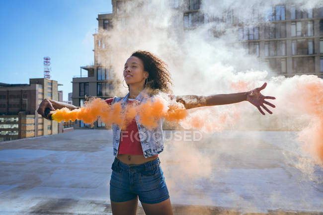 Vorderansicht einer jungen afrikanisch-amerikanischen Frau, die eine Jeansweste mit ausgestreckten Armen trägt und eine Rauchmaschine hält, die orangefarbenen Rauch auf einem Dach mit Blick auf ein Gebäude und Sonnenlicht produziert — Stockfoto