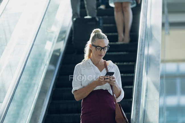 Vue de face de femme d'affaires caucasienne regardant le téléphone mobile tout en utilisant des escaliers mécaniques dans le bureau moderne — Photo de stock