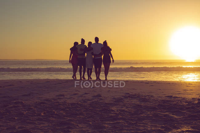 Вид ззаду на різноманітних друзів з гербами, що йдуть до пляжу під час заходу сонця — стокове фото