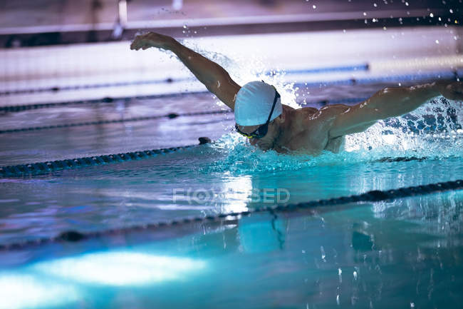 Вид сбоку на кавказского пловца в белой плавательной шапке и очках, делающих удар бабочки в бассейне — стоковое фото