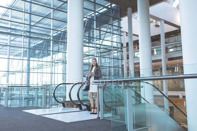Vue de face d'une femme d'affaires debout près d'un escalier roulant dans un immeuble de bureaux moderne — Photo de stock
