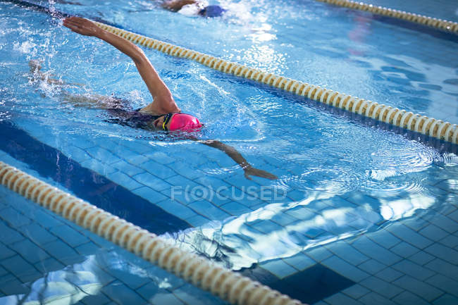 Vista lateral de uma jovem afro-americana e caucasiana fazendo freestyle acidente vascular cerebral na piscina enquanto o nadador com boné rosa leva — Fotografia de Stock