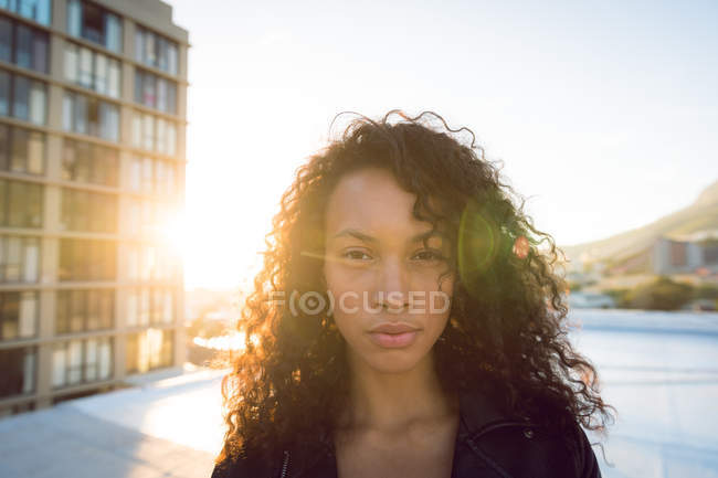 Close up of a young African-American woman wearing a leather jacket looking intently at the camera while standing on a rooftop with a view of a building and the sunset — Stock Photo