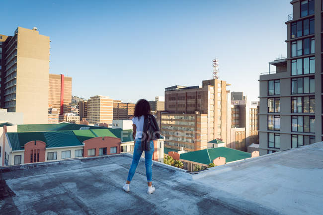 Vista posteriore di una giovane donna afro-americana con una giacca di pelle sopra la spalla in piedi su un tetto con vista sugli edifici — Foto stock