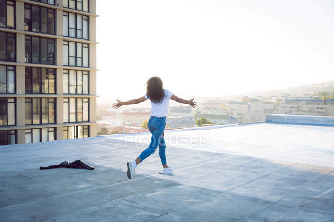 Вид сзади на молодую афроамериканку с протянутыми руками, бегущую по крыше с видом на здание и солнечный свет — стоковое фото