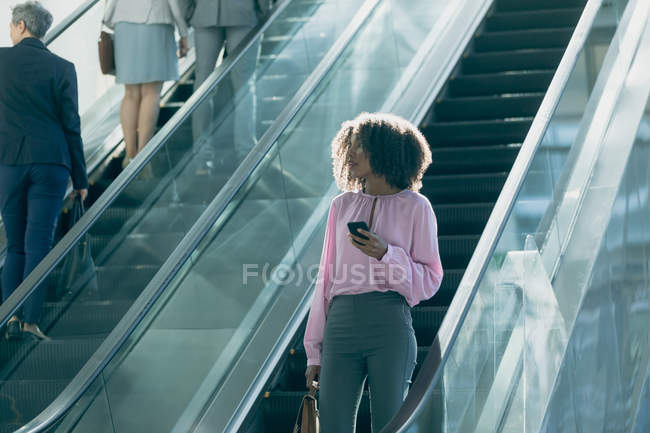 Vista frontal mujer de negocios afroamericana utilizando escaleras mecánicas en la oficina moderna - foto de stock