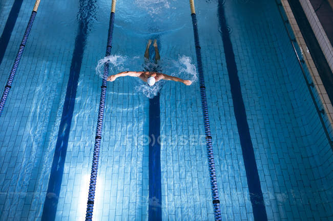 Высокий угол обзора самца белого пловца в белой плавательной шапочке, делающего инсульт бабочки в бассейне — стоковое фото