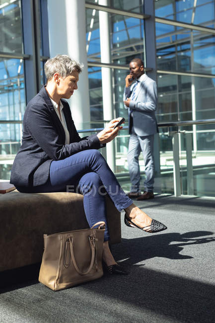 Side view of Caucasian female executive looking at her mobile phone while sitting on chair in modern office. Behind her, African american businessman speaking on mobile phone — Stock Photo