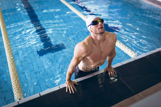 Vorderansicht eines männlichen kaukasischen Schwimmers mit weißer Badekappe und Schutzbrille, der den Körper seitlich des Schwimmbeckens nach oben hebt — Stockfoto