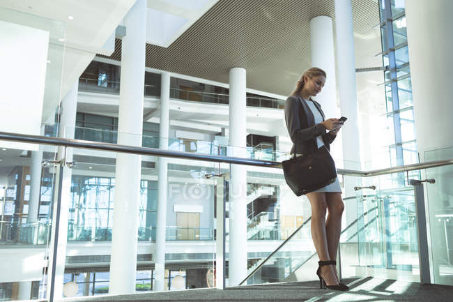 Низький кут зору бізнес-леді, спираючись на перила і використовуючи мобільний телефон в сучасній офісній будівлі — стокове фото