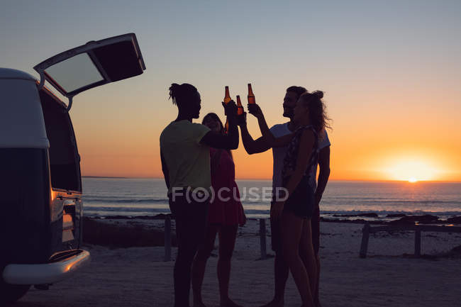 Vista lateral do grupo de amigos diversos brindar garrafas de cerveja perto van campista durante o pôr do sol — Fotografia de Stock