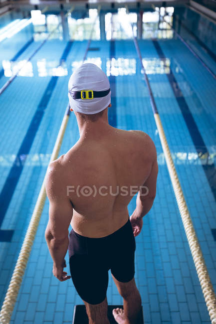 Rear view of a male Caucasian swimmer wearing a white swimming cap standing by an olympic sized pool inside a stadium — Stock Photo
