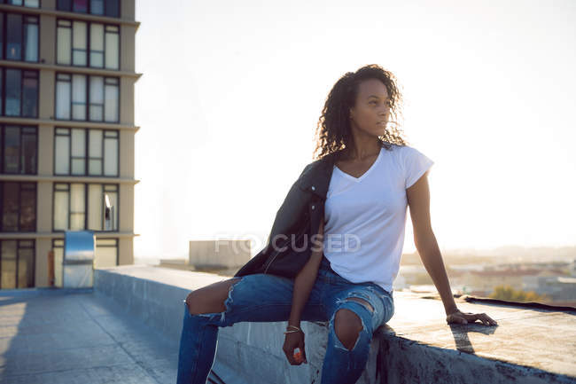 Vista frontal de una joven afroamericana con una chaqueta de cuero sobre el hombro sentada mientras mira lejos de la cámara en una azotea con una vista de un edificio y la luz del sol - foto de stock