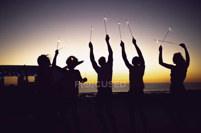 Vista frontal de la silueta de diversos amigos jugando con bengalas en la playa al atardecer - foto de stock