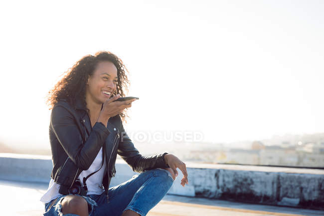 Вид спереди на молодую афроамериканку в кожаной куртке, улыбающуюся при использовании мобильного телефона и сидящую на крыше — стоковое фото