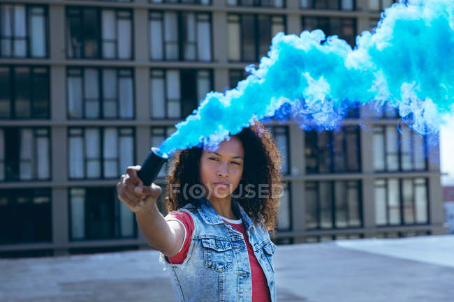 Vista frontal de una joven afroamericana vestida con un chaleco vaquero sosteniendo un fabricante de humo que produce humo azul en una azotea con vista a un edificio - foto de stock