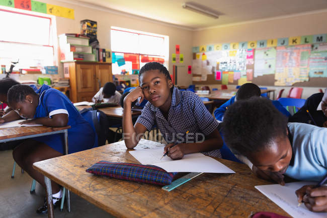 Front view of a young African schoolgirl leaning on her desk and looking up while writing in her notebook during a lesson in a township elementary school classroom, beside her and in the background her classmates are writing in their books — Stock Photo