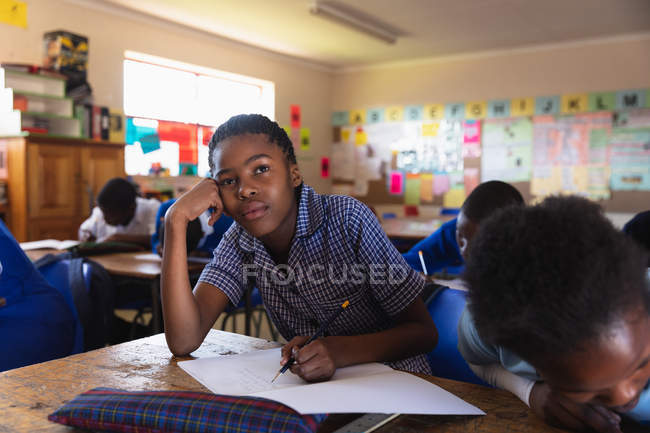 Front view close up of a young African schoolgirl leaning on her desk and looking up while writing in her notebook during a lesson in a township elementary school classroom, in the background her classmates are also writing in their books — Stock Photo