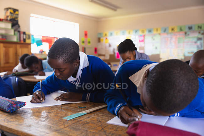 Vista frontal de perto de dois jovens meninos africanos escrevendo em seus cadernos durante uma aula em uma sala de aula da escola primária da cidade, em segundo plano seus colegas também estão escrevendo em seus livros — Fotografia de Stock