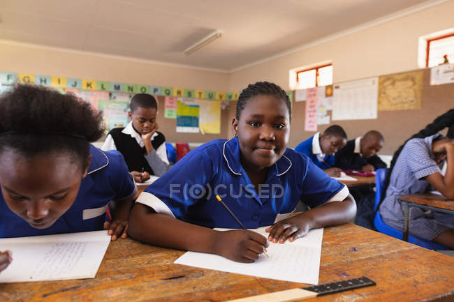 Portrait close up of a young African schoolgirl leaning on her desk, looking to camera and smiling while writing in her notebook during a lesson in a township elementary school classroom. — Stock Photo
