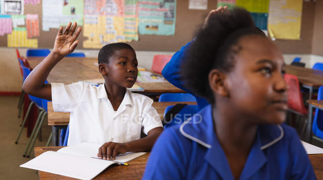 Vista frontal de un joven escolar africano sentado en su escritorio, levantando la mano para responder a una pregunta durante una lección en un aula de la escuela primaria del municipio, en primer plano una colegiala está escuchando atentamente - foto de stock