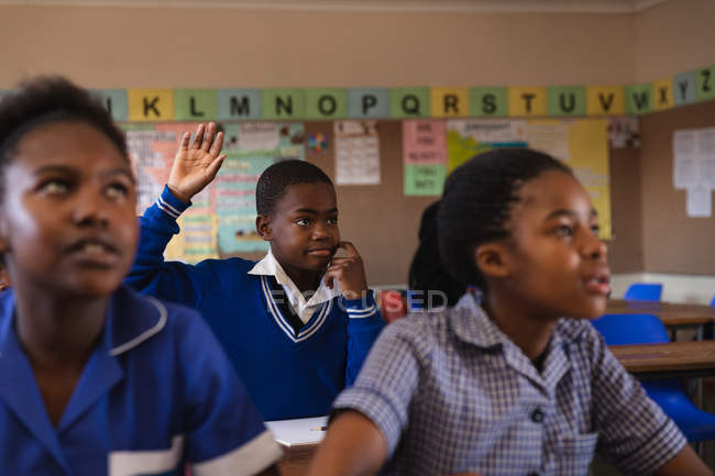 Вид спереду молодий Африканський школяр сидить на своєму столі, піднявши руку, щоб відповісти на питання під час уроку в містечку початкової школи школі, на передньому плані два школярки слухають уважно — стокове фото
