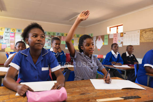 Frontansicht von zwei jungen afrikanischen Schulmädchen, die an ihrem Schreibtisch sitzen, eine hebt ihre Hand, um während einer Schulstunde in einer Township-Grundschule eine Frage zu beantworten, im Hintergrund hören ihre Mitschüler aufmerksam zu. — Stockfoto