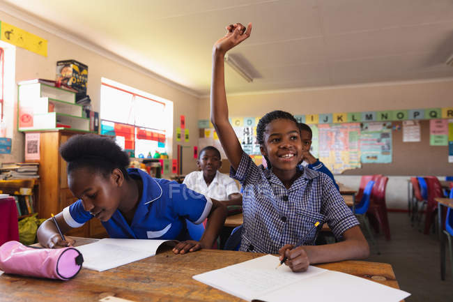 Frontansicht von zwei jungen afrikanischen Schulmädchen, die an ihrem Schreibtisch sitzen, wobei eine ihre Hand hebt, um eine Frage zu beantworten, und die andere während einer Unterrichtsstunde in ihr Notizbuch schreibt. — Stockfoto