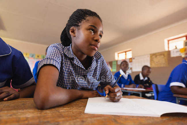 Front view close up of a young African schoolgirl sitting at her desk and looking up while writing in her notebook during a lesson — Stock Photo