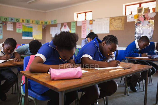 Vista frontal de cerca de un grupo de jóvenes escolares africanos que escriben en sus cuadernos durante una lección en un aula de la escuela primaria del municipio - foto de stock