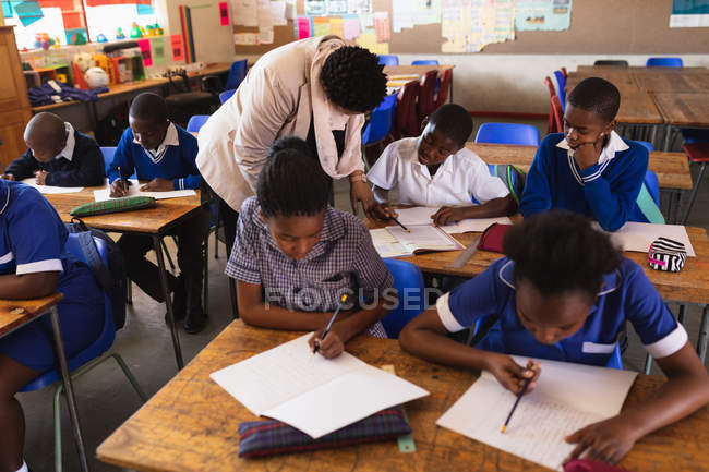 Vista elevada de una maestra de escuela africana de mediana edad ayudando a dos jóvenes escolares africanos sentados en su escritorio durante una lección en un aula de escuela primaria del municipio, mientras que sus compañeros de clase están ocupados leyendo y escribiendo en sus libros - foto de stock