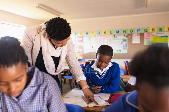 Close up front view of a middle aged African female school teacher helping a young African schoolboy sitting at his desk during a lesson in a township elementary school classroom, while classmates are busy reading and writing in their books — Stock Photo