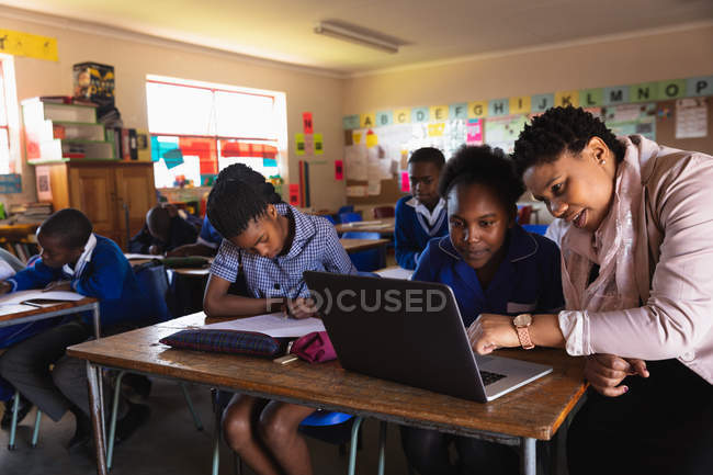Front view close up of a middle aged African female school teacher helping a young African schoolgirl sitting at her desk using a laptop computer during a lesson. — Stock Photo