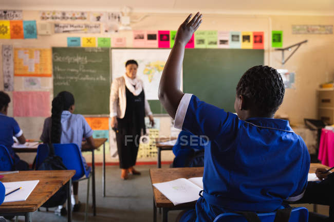 Задний вид молодой африканской школьницы, поднимающей руку, чтобы ответить на вопрос учительнице, стоящей перед классом во время урока в городском классе начальной школы — стоковое фото