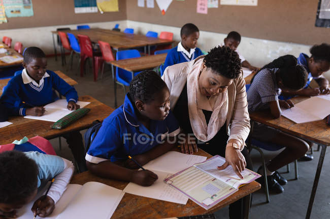 Elevated view of a middle aged African female school teacher helping a young African schoolgirl sitting at her desk during a lesson — Stock Photo