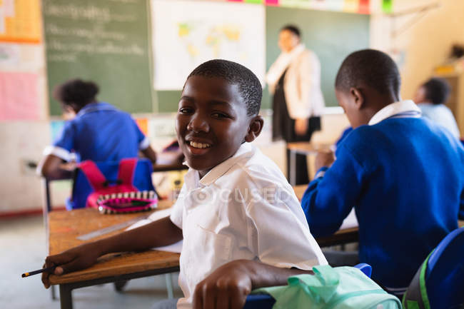 Side view close up of a young African schoolboy sitting at his desk and turning around, looking to camera and smiling during a lesson in a township elementary school classroom. — Stock Photo