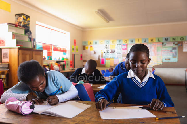 Вид спереди на молодую африканскую школьницу, сидящую за своим столом и пишущую в своей книге, и молодого африканского школьника, сидящего рядом с ней, смотрящего вниз и думающего во время урока — стоковое фото