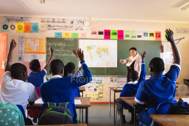 Вид сзади на молодых африканских школьников, поднимающих руки, чтобы ответить на вопрос учительнице, стоящей перед классом у доски во время урока в классе начальной школы. — стоковое фото