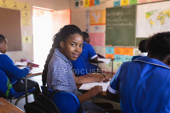 Side view close up of a young African schoolgirl sitting at her desk and turning around, looking to camera and smiling during a lesson in a township elementary school classroom. — Stock Photo