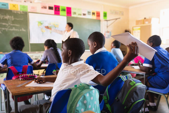 Vista laterale da vicino di un giovane scolaro africano seduto alla scrivania e che lancia un aeroplano di carta, durante una lezione in una classe della scuola elementare cittadina . — Foto stock