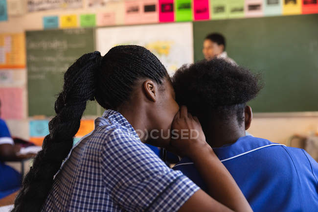 Visão traseira close-up de duas jovens alunas africanas sentadas em sua mesa sussurrando umas às outras durante uma aula em uma sala de aula da escola primária da cidade — Fotografia de Stock
