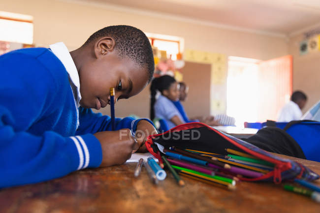 Vista lateral de perto de um jovem estudante africano sentado em sua mesa escrevendo durante uma aula em uma sala de aula da escola primária da cidade, em segundo plano os colegas também estão sentados em suas mesas escrevendo — Fotografia de Stock