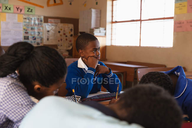 Side view of a young African schoolboy sitting at his desk, leaning and listening during a lesson in a township elementary school classroom, around him classmates are sitting at their desks writing — Stock Photo