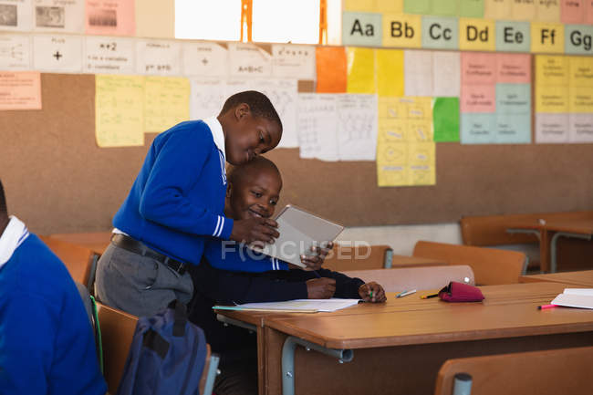 Front view of two young African schoolboys looking at a tablet computer and talking during a lesson in a township elementary school classroom. — Stock Photo