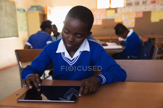 Front view close up of a young African schoolboy sitting at his desk using a tablet computer and smiling during a lesson in a township elementary school classroom, in the background classmates are sitting at their desks working — Stock Photo