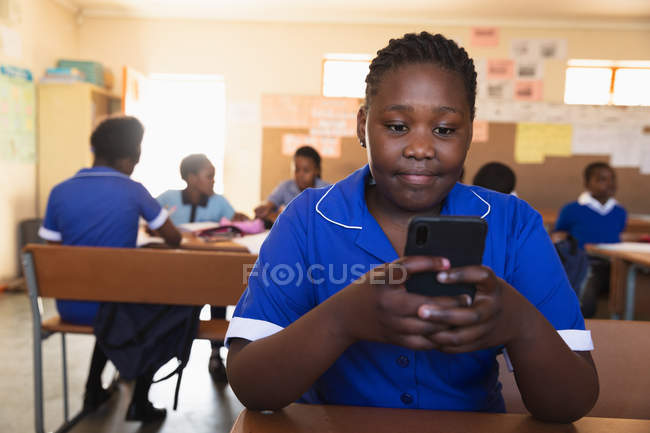 Vue de face gros plan d'une jeune écolière africaine assise à son bureau à l'aide d'un smartphone et souriant dans une salle de classe d'une école élémentaire d'un canton . — Photo de stock