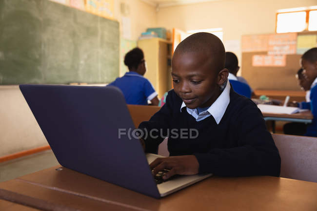 Vista frontal de cerca de un joven colegial africano sentado en su escritorio usando una computadora portátil y sonriendo durante una lección en un aula de la escuela primaria del municipio, en el fondo los compañeros de clase están sentados en sus escritorios trabajando - foto de stock