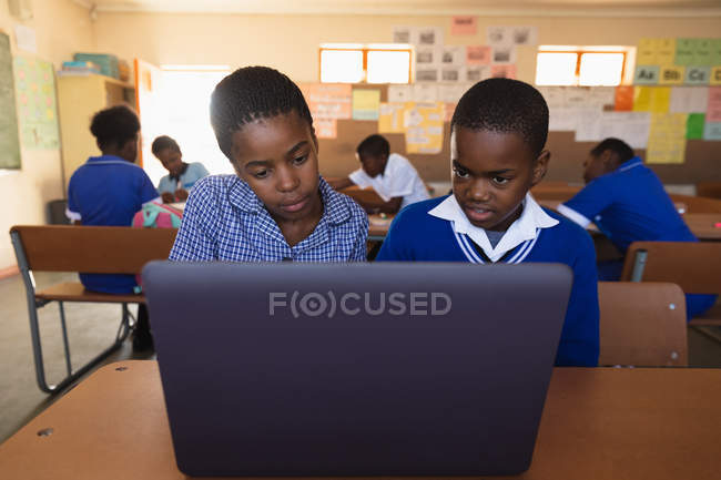 Vue de face gros plan d'un jeune écolier africain et d'une écolière assis à un bureau à l'aide d'un ordinateur portable ensemble pendant une leçon dans une classe de l'école élémentaire d'un canton, en arrière-plan, des camarades de classe sont assis à leur bureau travaillant — Photo de stock