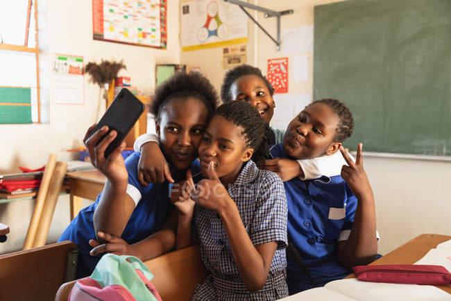 Vista frontal close-up de um grupo de jovens alunas africanas se divertindo posando e tirando selfies com um smartphone durante uma pausa das aulas em uma sala de aula da escola primária da cidade — Fotografia de Stock